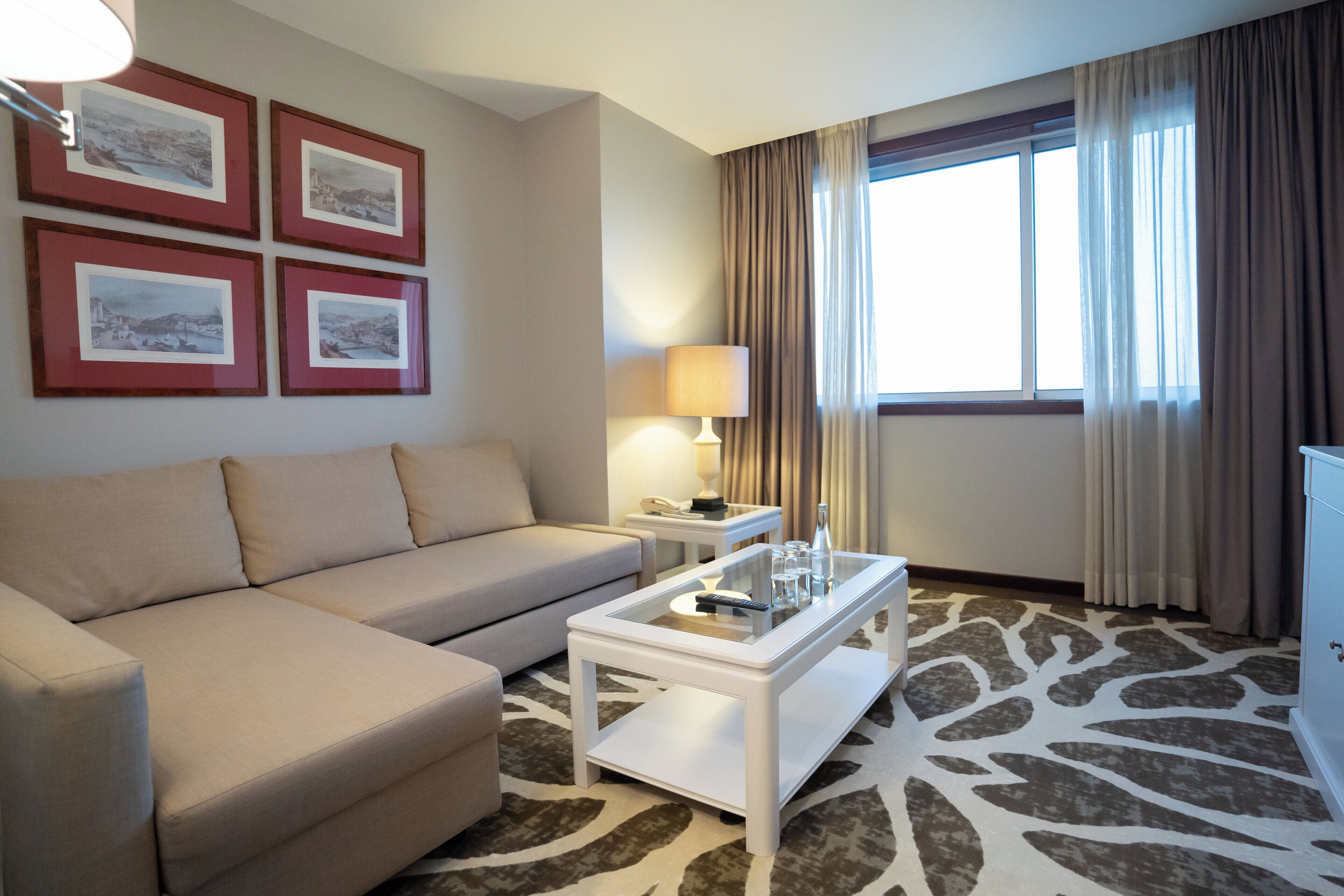 A hotel room with a beige sofa, a glass-top coffee table, a lamp, and framed pictures on the wall.