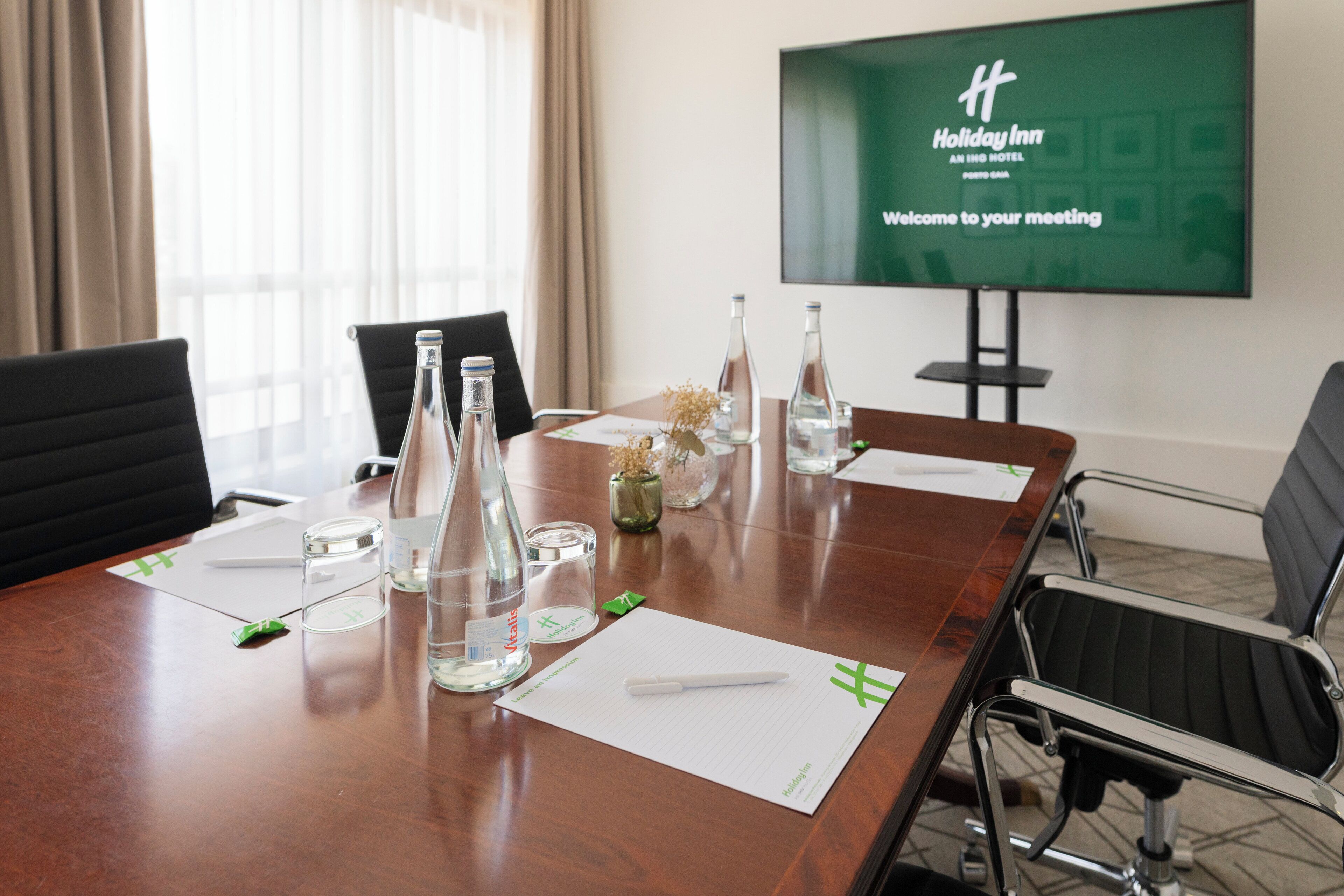 A conference room with a wooden table, chairs, a projector screen displaying a welcome message, and a window with curtains.