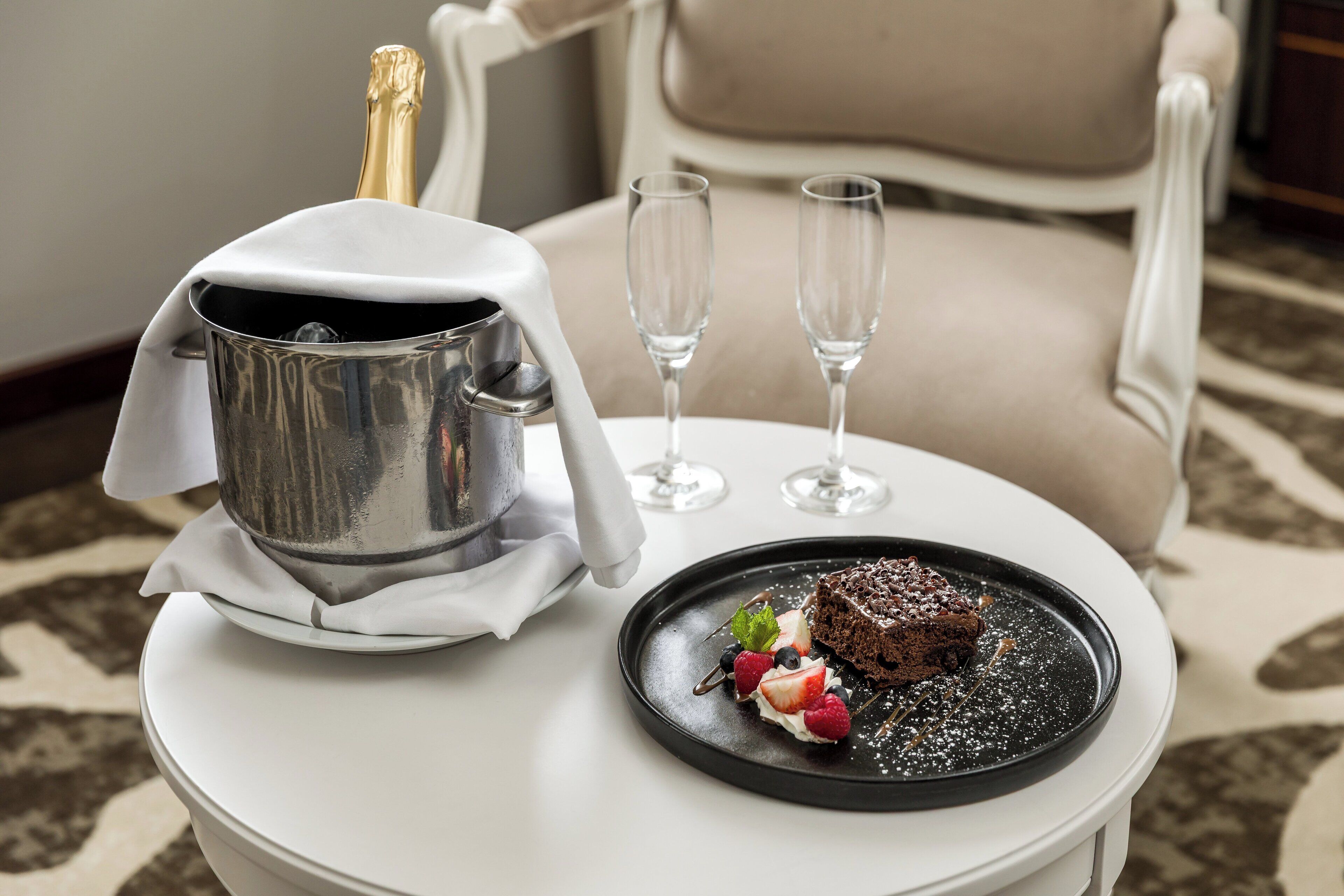 A dessert plate with chocolate cake and berries on a white table, a champagne bottle, and two empty wine glasses.
