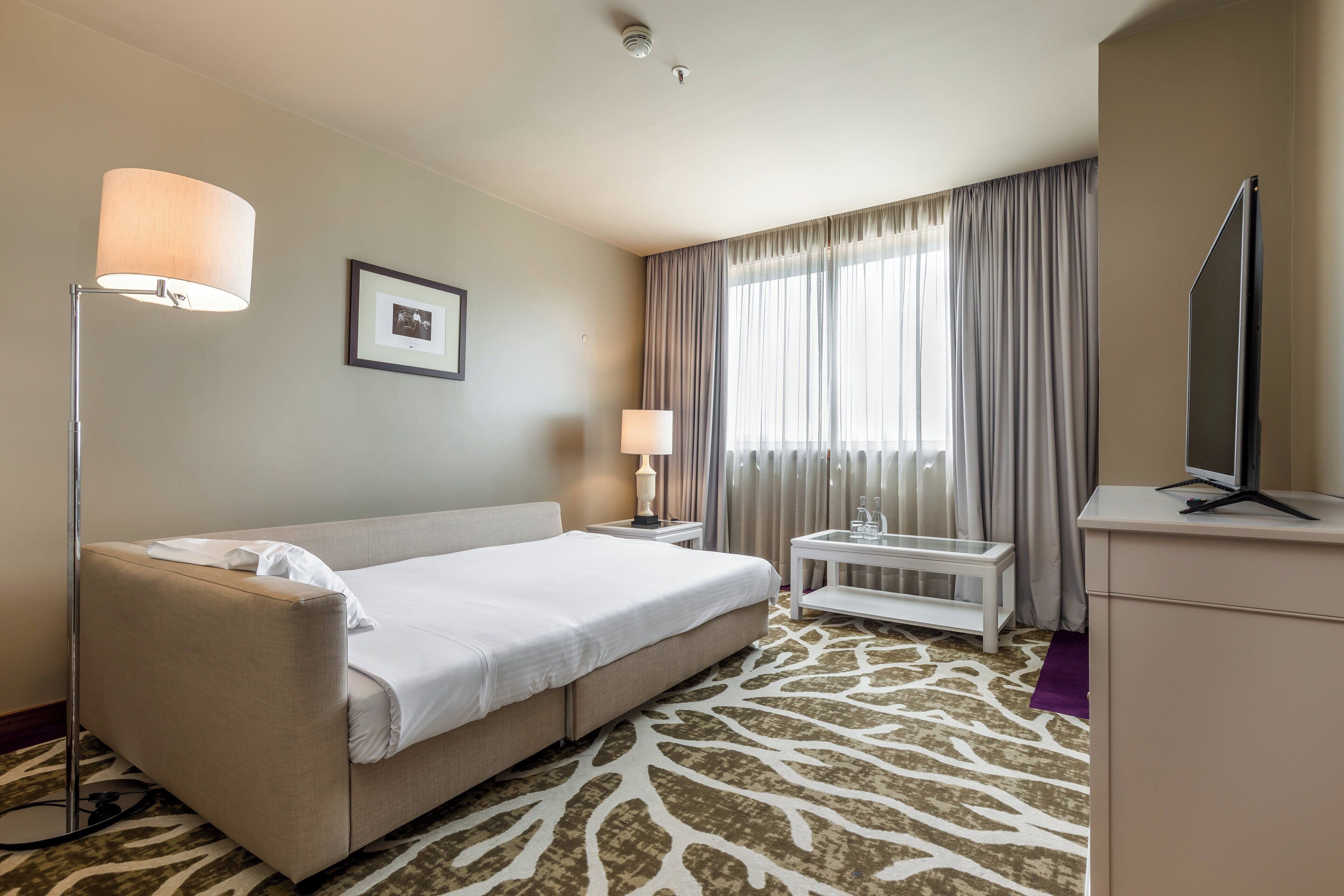 A hotel room with a bed, a desk, a lamp, a television, and a window with curtains.