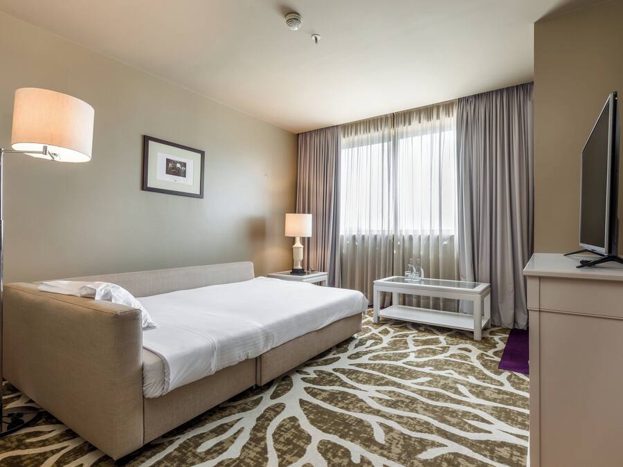 A hotel room with a bed, a desk, a lamp, a television, and a window with curtains.
