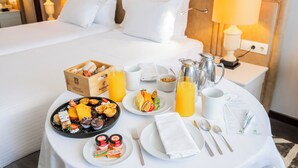 Een net gedekte ontbijttafel met diverse gebakjes, fruit en drankjes, in een hotelkamer met een groot bed en nachtlampjes.