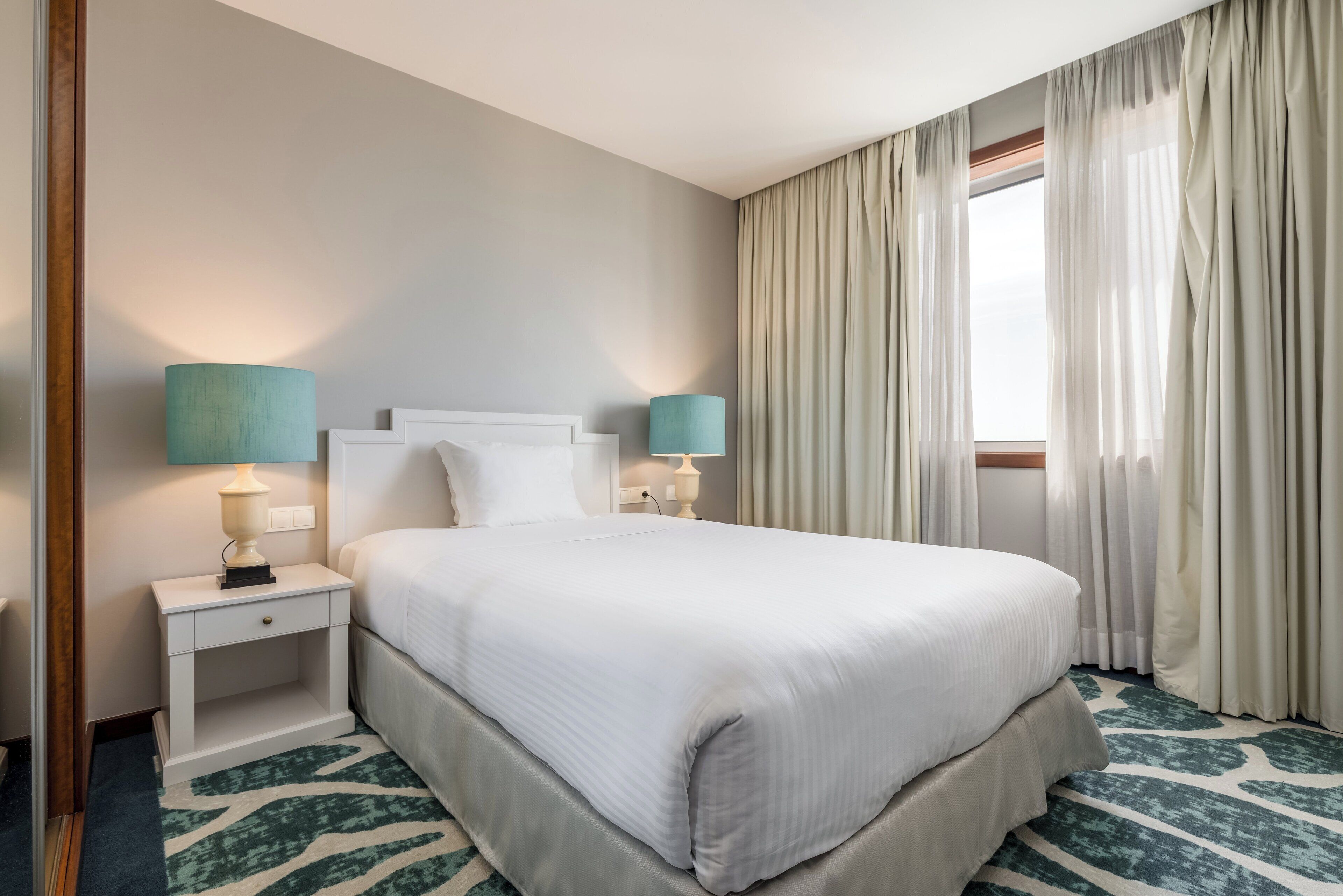 Hotellihuone, jossa on suuri sänky, kaksi yöpöytää valaisimineen, ikkuna verhoineen ja kuviollinen matto.