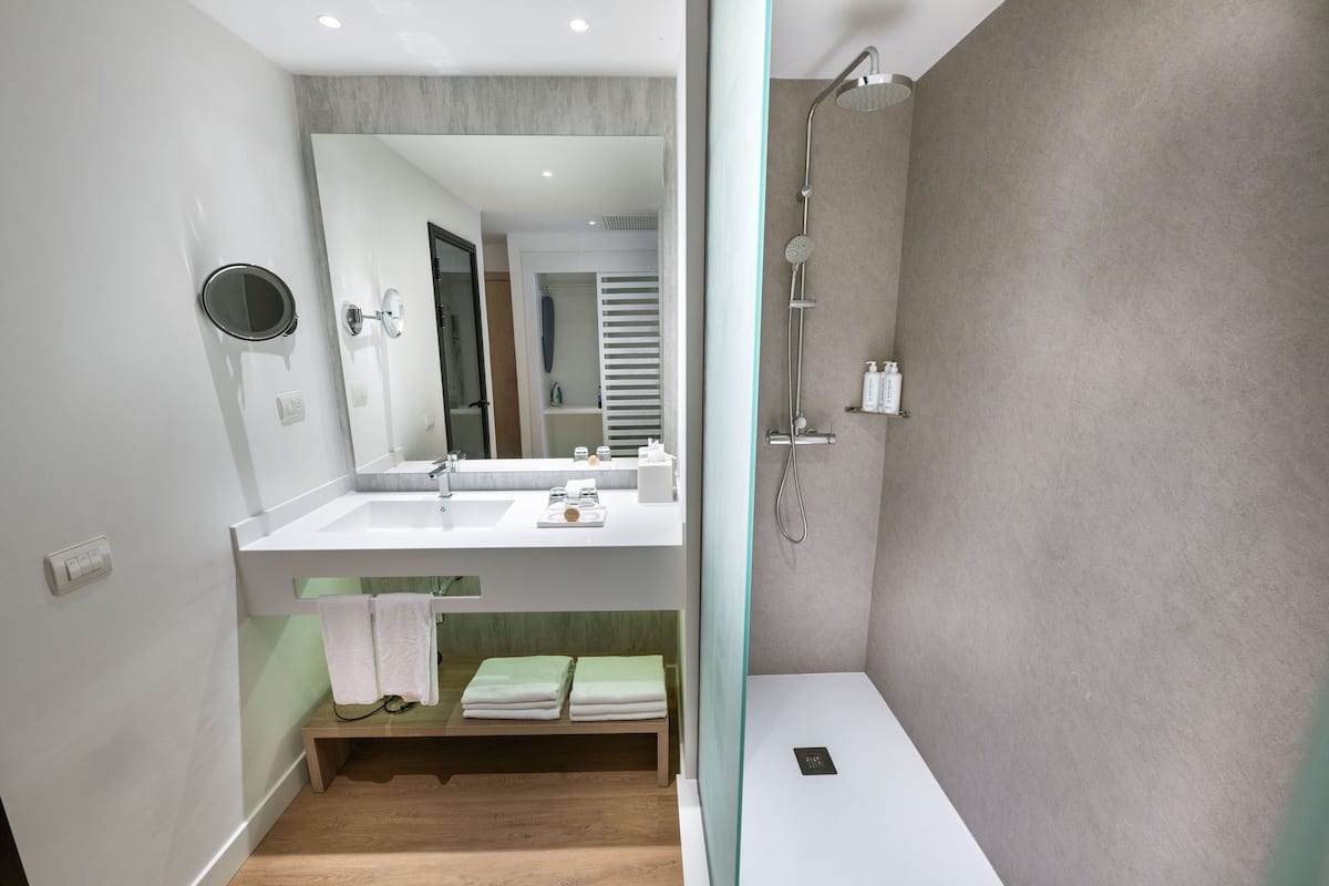 Un baño moderno con ducha, lavabo y espejo. Hay un banco con toallas y un teléfono inteligente encima.