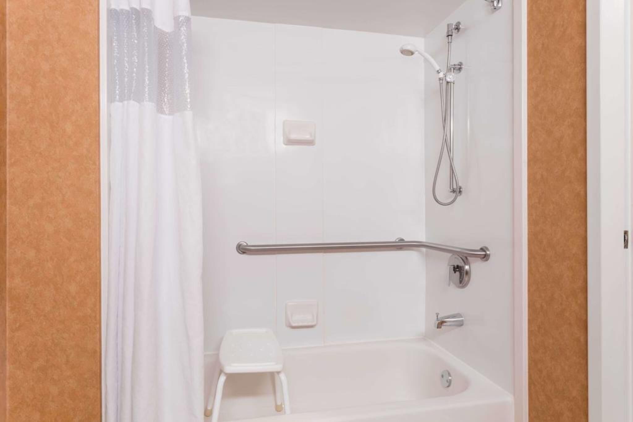 Un baño con bañera, ducha, barra de apoyo, taburete pequeño y jabonera.
