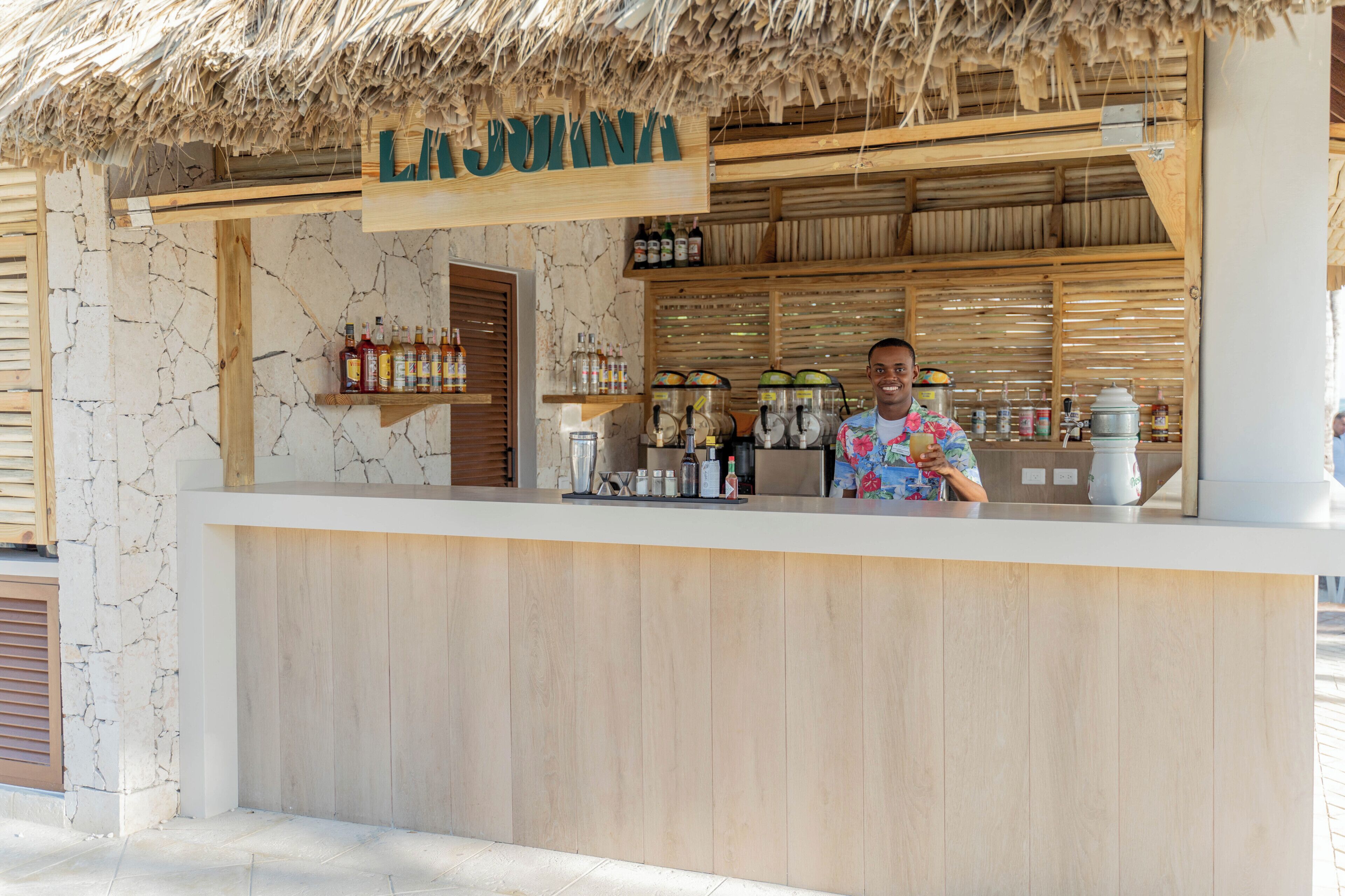 A bar area with a wooden counter, a sign above it, and shelves with bottles.