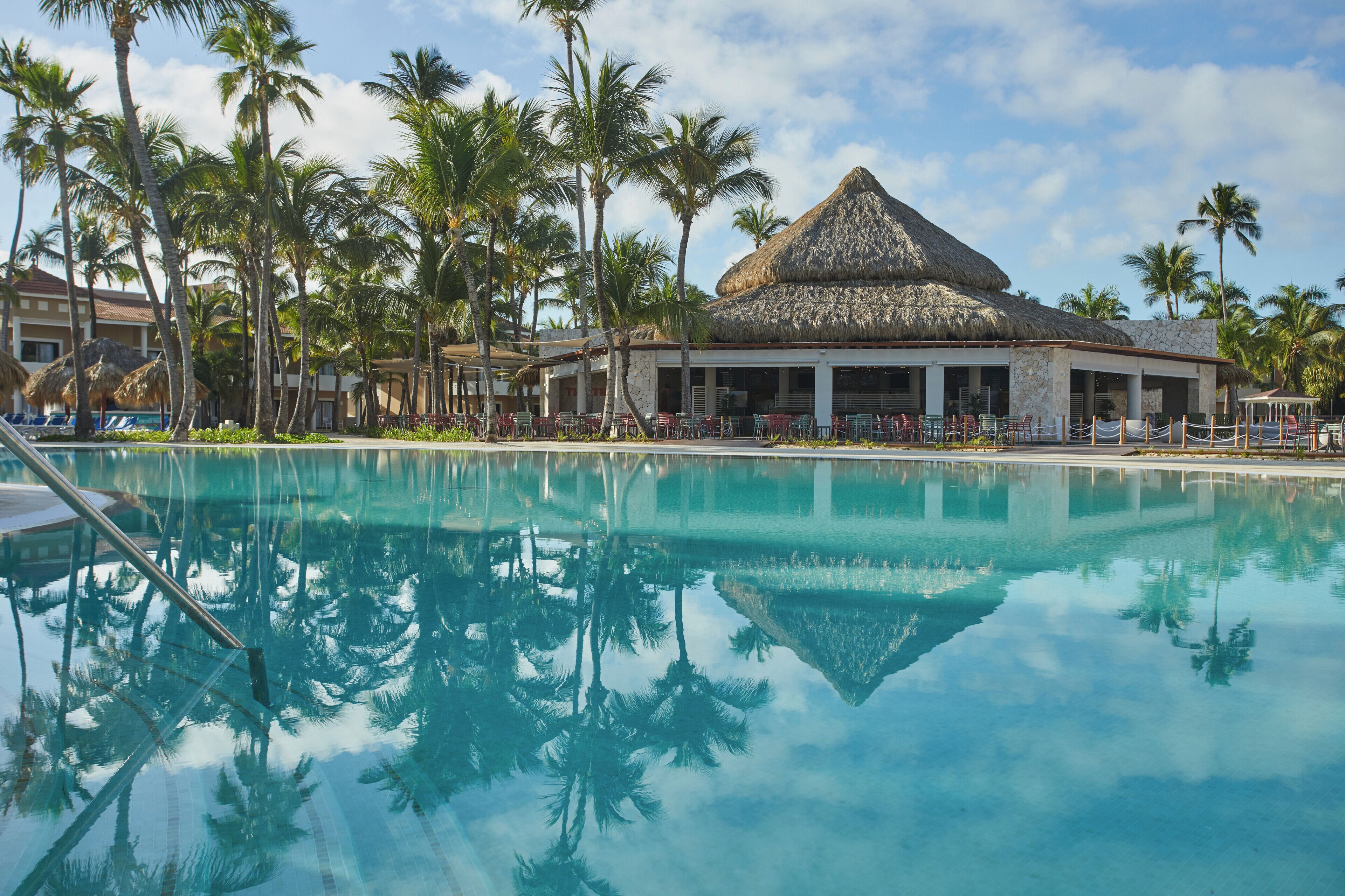 A resort with a pool, palm trees, and a thatched-roof building.