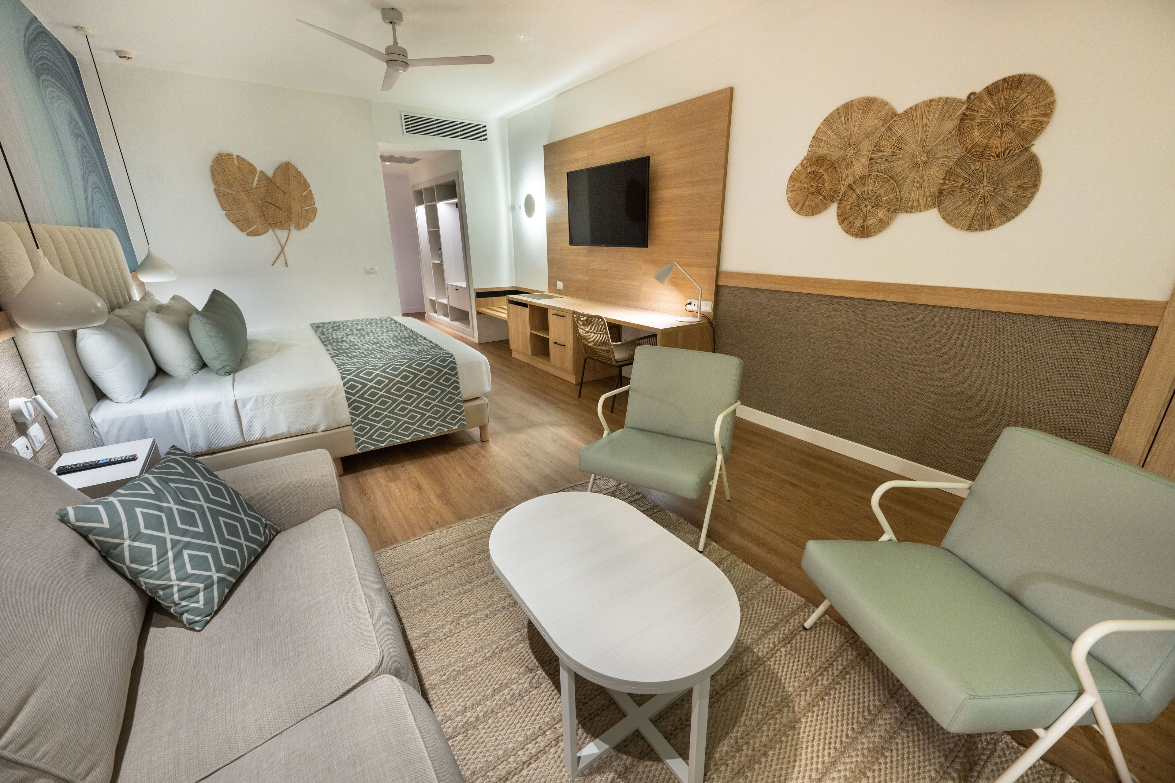 A modern hotel room with a sofa, armchair, and a small table. There is a desk with a chair and a flat-screen TV mounted on the wall.