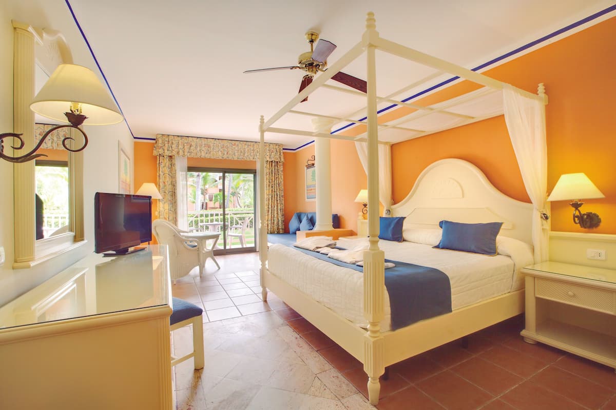 A four-poster bed with a canopy, a television, a desk with a chair, and a balcony with a view of palm trees.