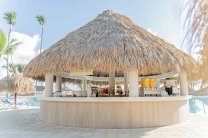 Zona bar circolare con tetto di paglia, circondata da palme e una piscina.