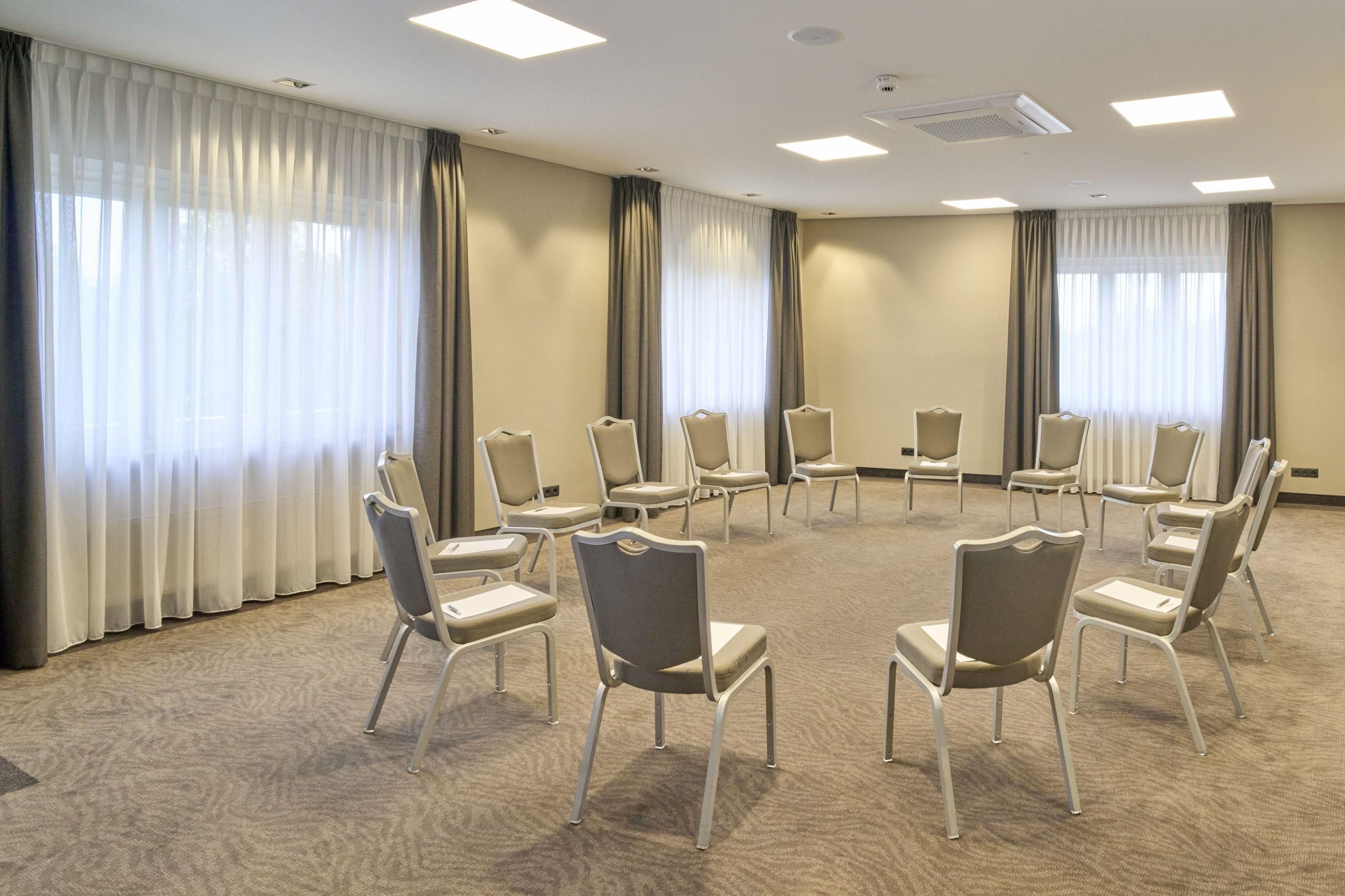 Un salón de conferencias con sillas dispuestas en círculo, alfombra color beige y grandes ventanales con cortinas.