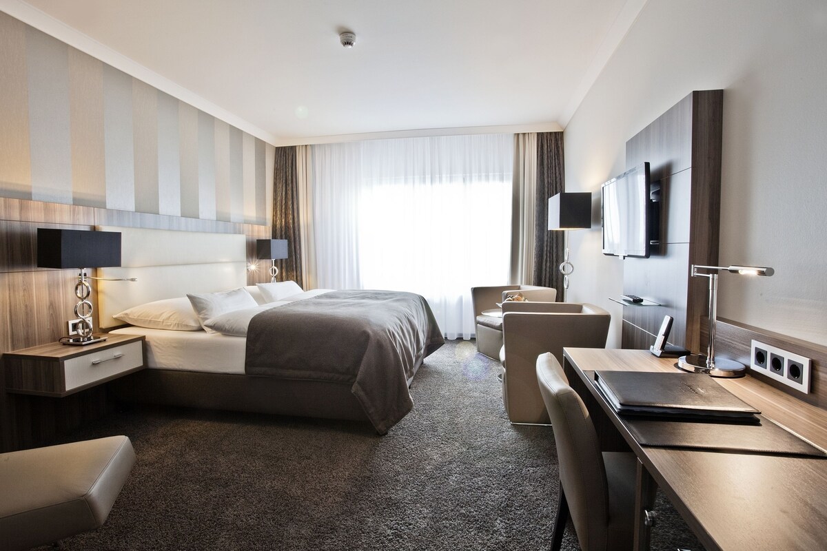 Une chambre d’hôtel moderne équipée d’un grand lit, d’un bureau, d’une chaise, d’une télévision et d’une fenêtre avec des rideaux.