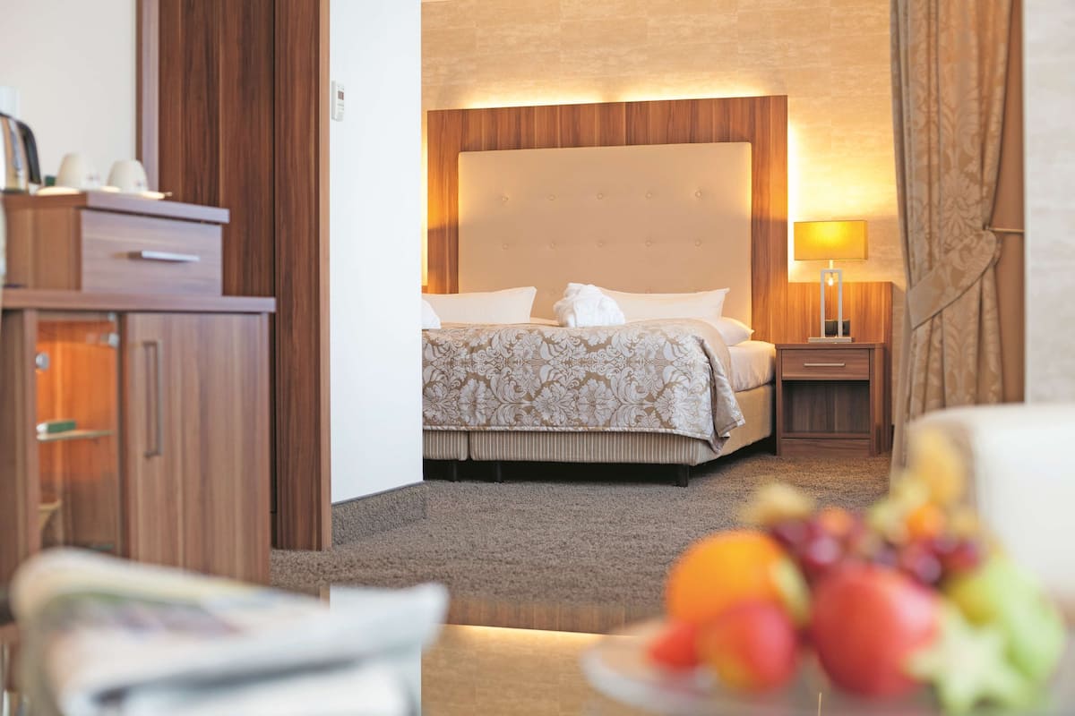 Quarto de hotel com cama grande, cabeceira de madeira, mesa de cabeceira com abajur e um pequeno armário.