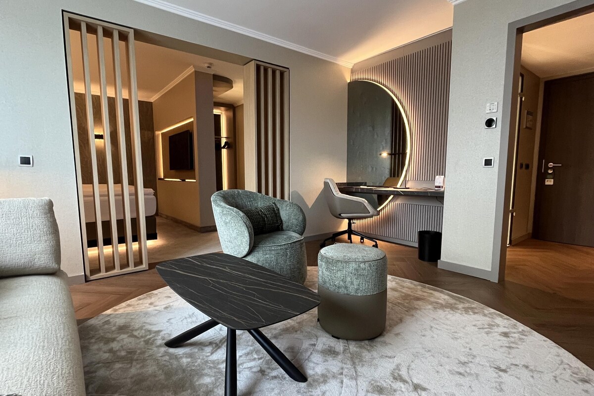 Een moderne woonkamer met een bank, een salontafel en een stoel. Er hangt een spiegel aan de muur en een deur die leidt naar een andere kamer.