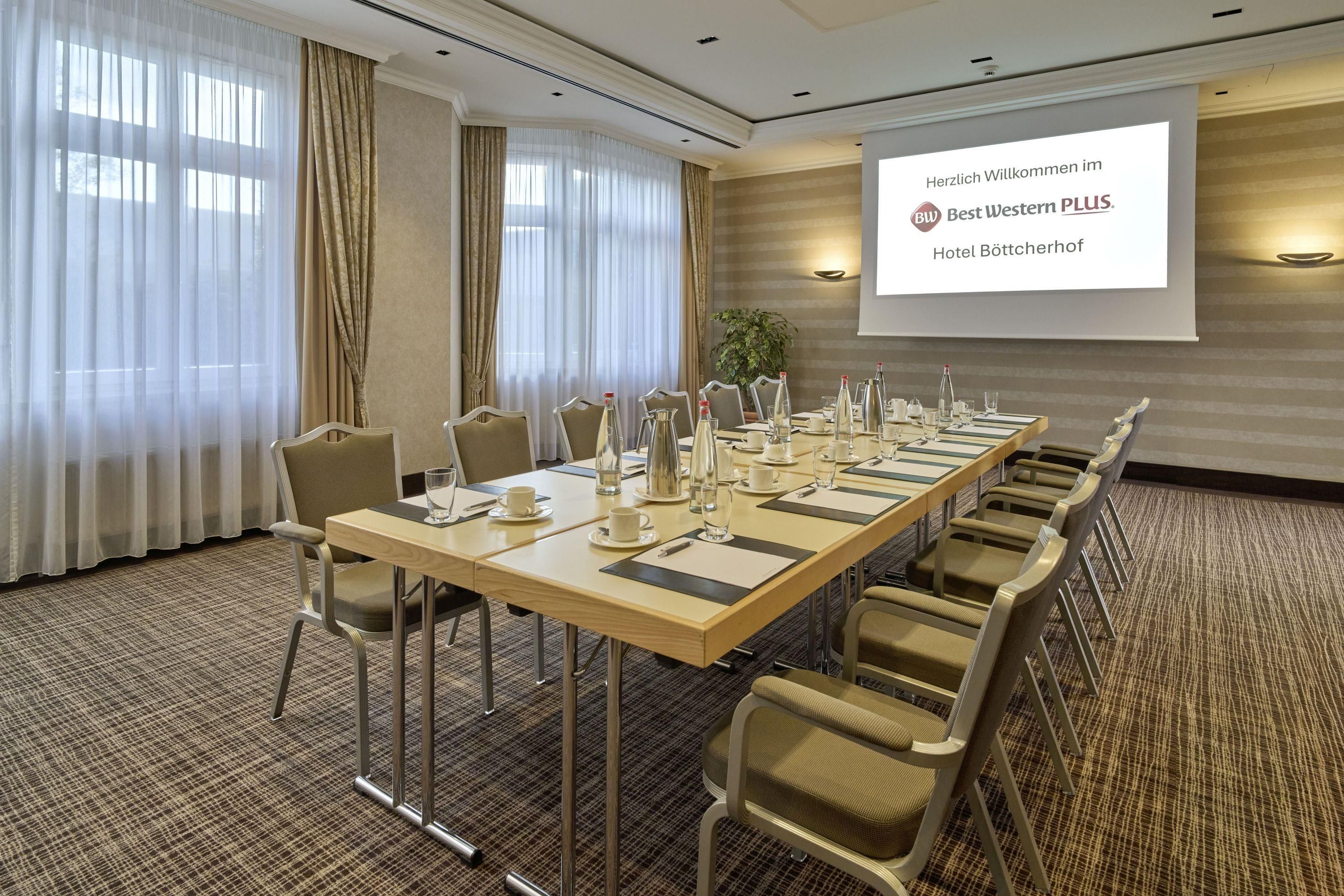 A conference room with a long table set for meetings, equipped with chairs and a projector screen displaying a welcome message.
