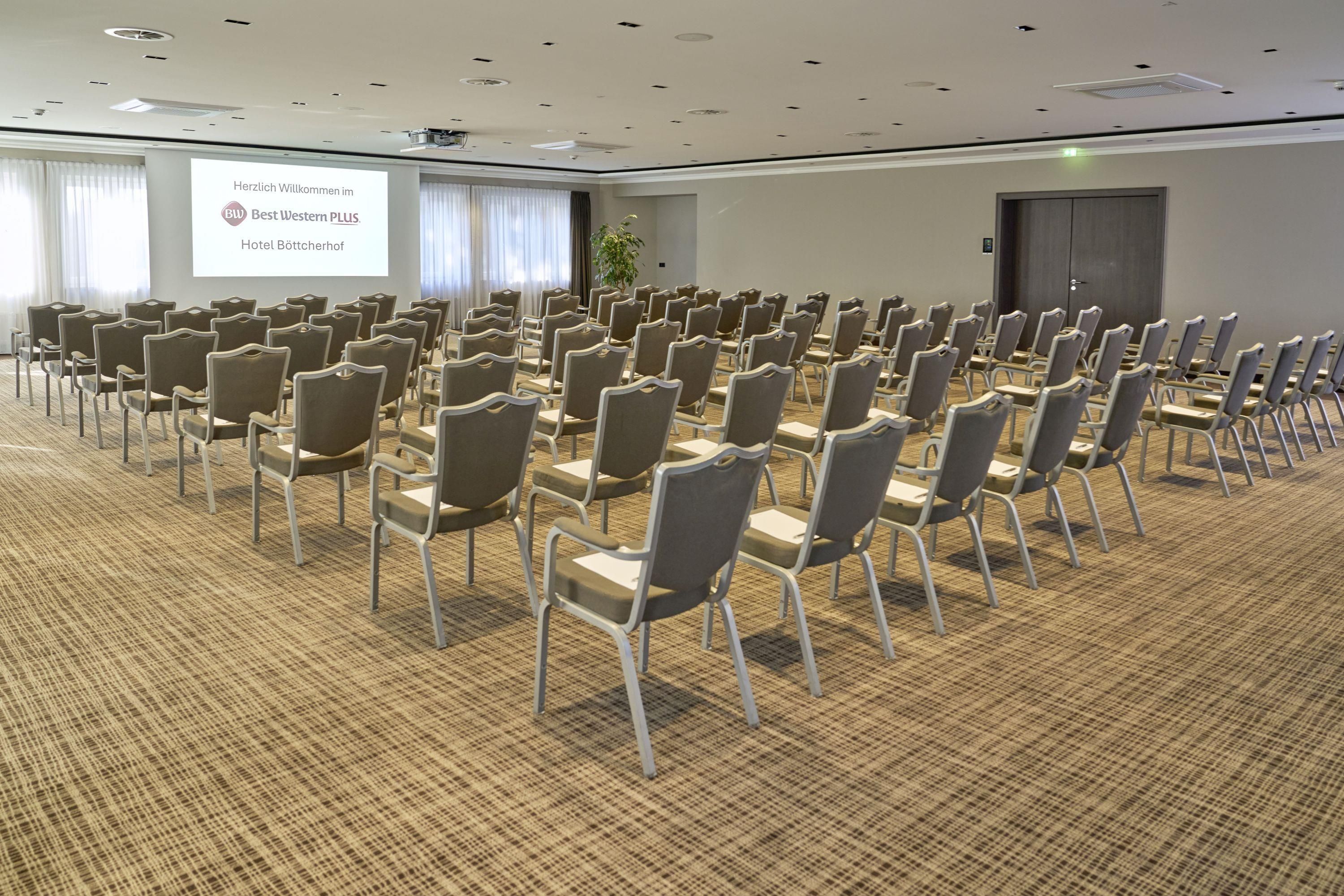 Un salón de conferencias con filas de sillas, una pantalla de proyector y piso alfombrado.