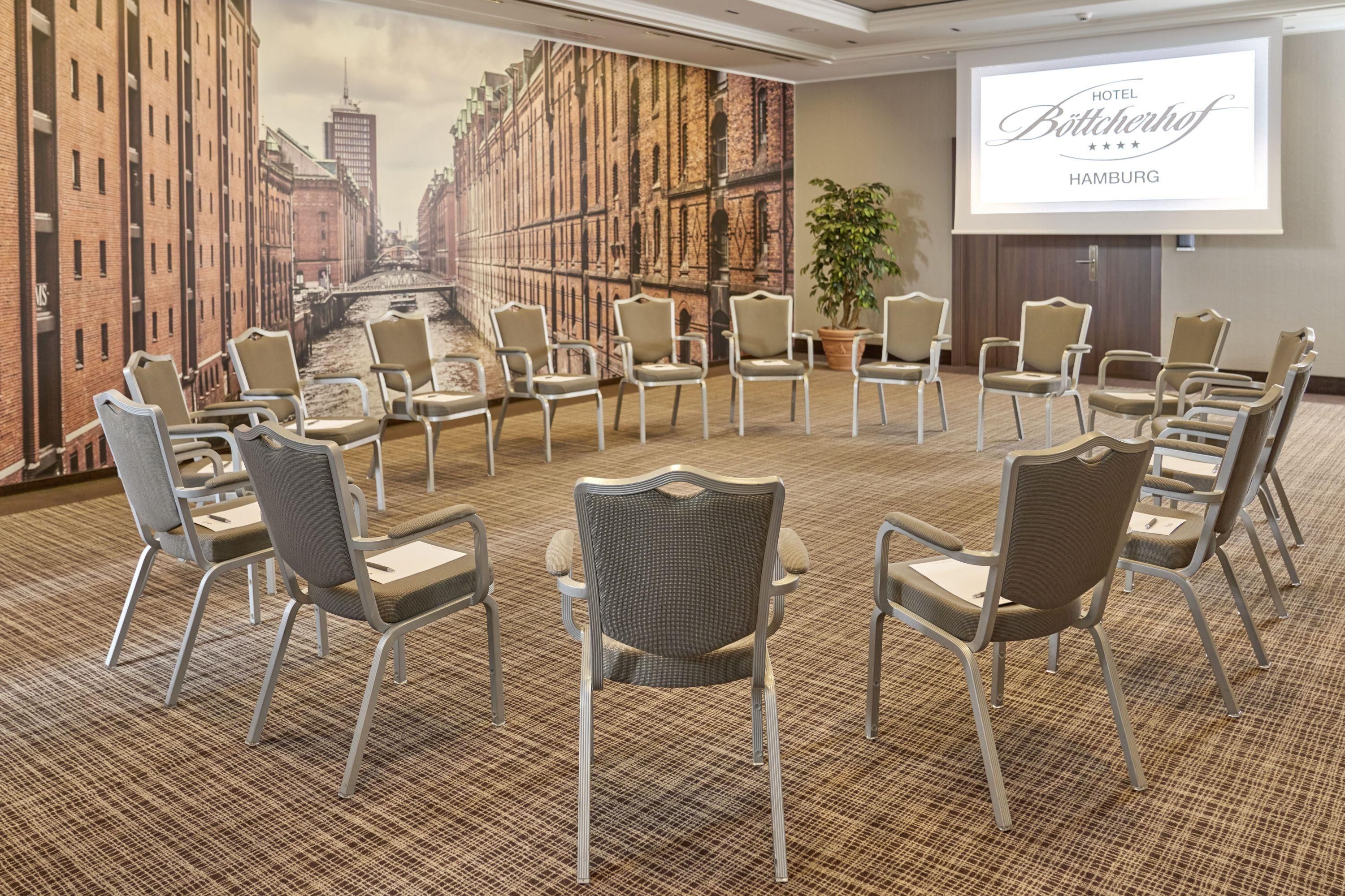 Une salle de conférence ornée d’une fresque représentant un paysage urbain, avec des chaises disposées en cercle et un écran de projection affichant le texte « HÔTEL Böttcherhof HAMBOURG ».