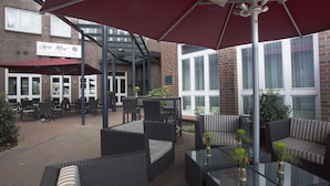 Un espace extérieur aménagé avec des meubles en osier et des tables en verre, un bâtiment en briques sur lequel on peut lire l’enseigne d’un salon de dégustation de cigares, et de grandes fenêtres.