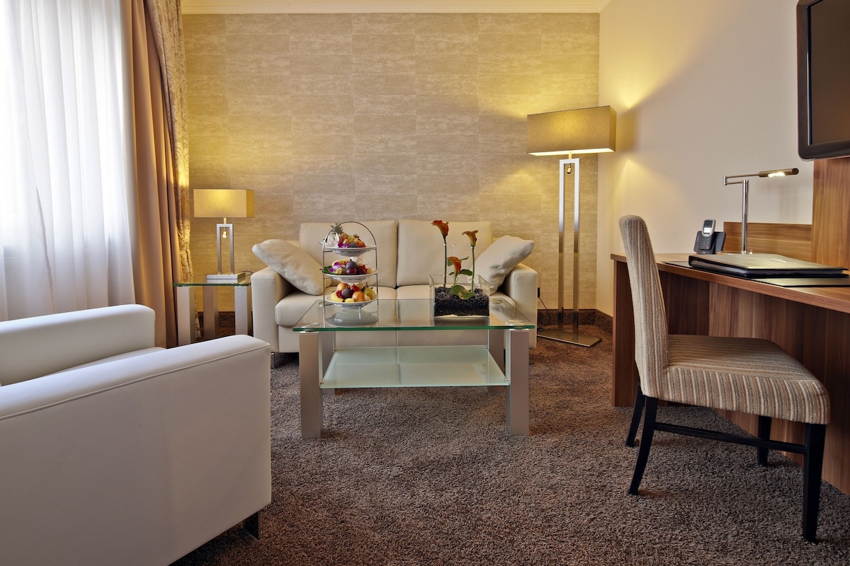 Une chambre d’hôtel comprenant un canapé, une table basse en verre avec des fruits, une table d’appoint avec une lampe, un bureau avec une chaise et un téléviseur fixé au mur.