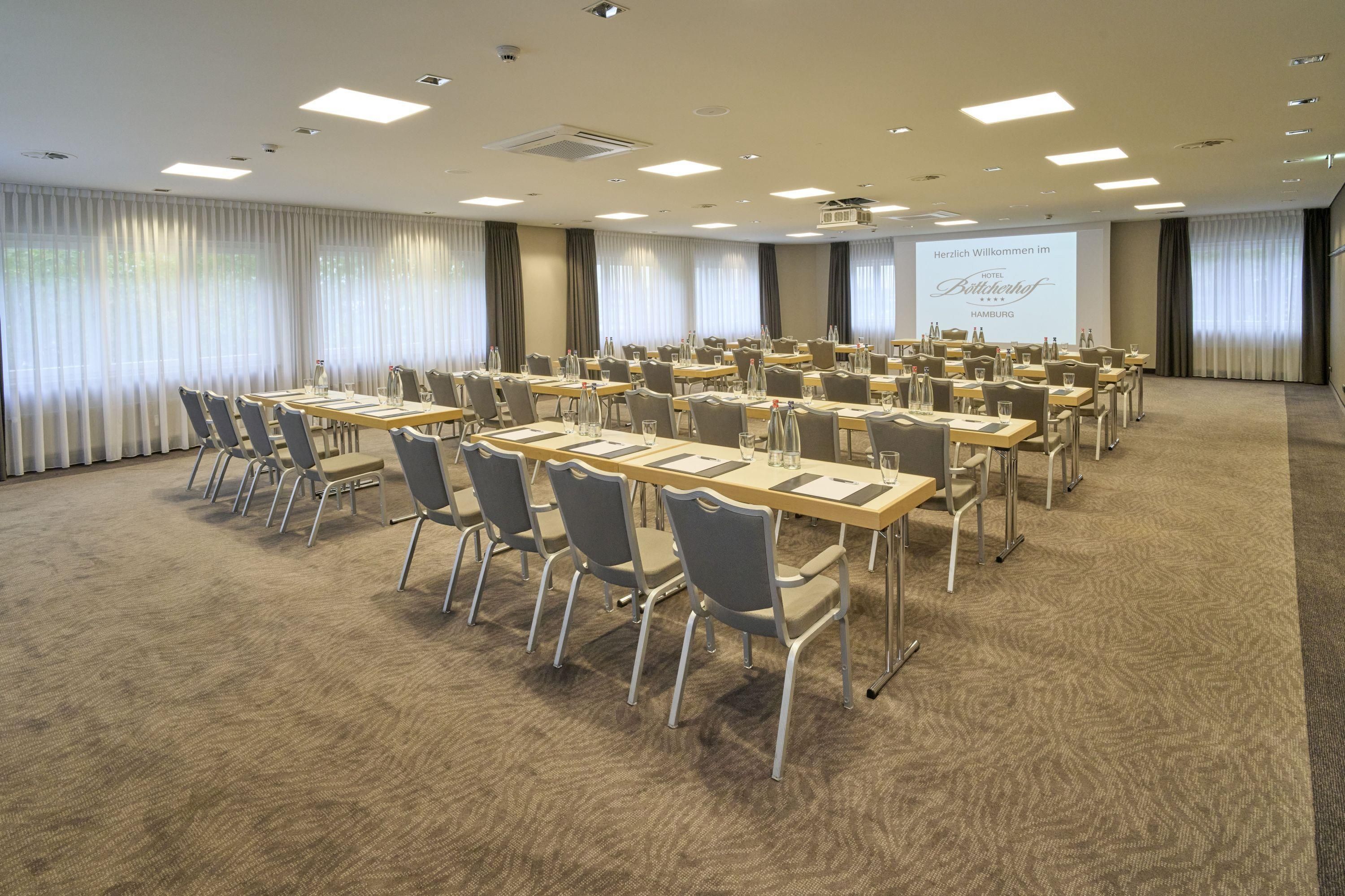 A conference room with rows of tables and chairs, a projector screen, and a carpeted floor.