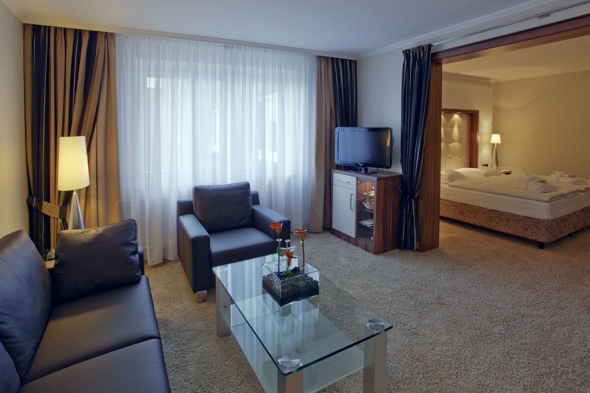 Une chambre d’hôtel avec un canapé, une table basse en verre, un téléviseur, un lit et une salle de bain visible à travers une porte ouverte.