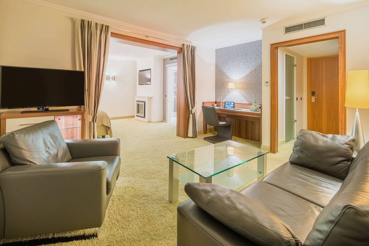 Quarto de hotel com sofá de couro, mesa de centro de vidro, televisão de tela plana, escrivaninha com computador e abajur.
