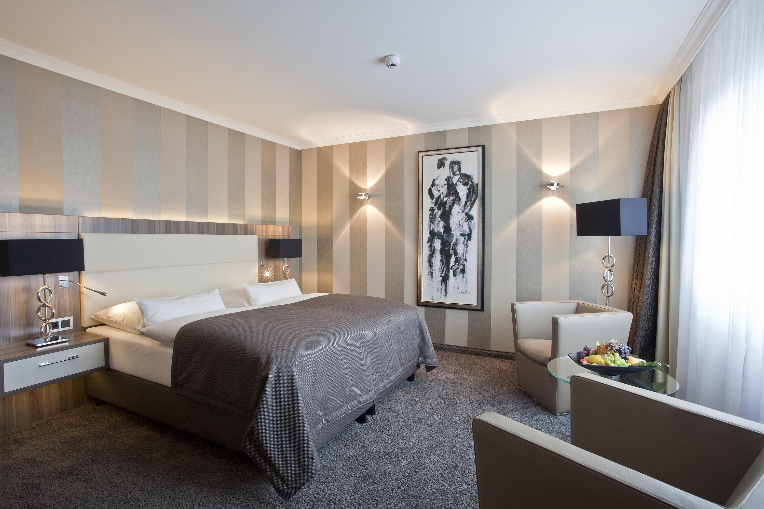 Habitación de hotel con una cama grande, lámparas de noche, una obra de arte en la pared, una mesita con fruta y una zona de estar.