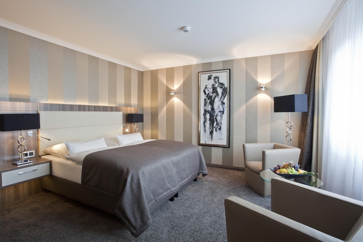 Hotelkamer met een groot bed, bedlampen, een kunstwerk aan de muur, een klein tafeltje met fruit en een zithoek.