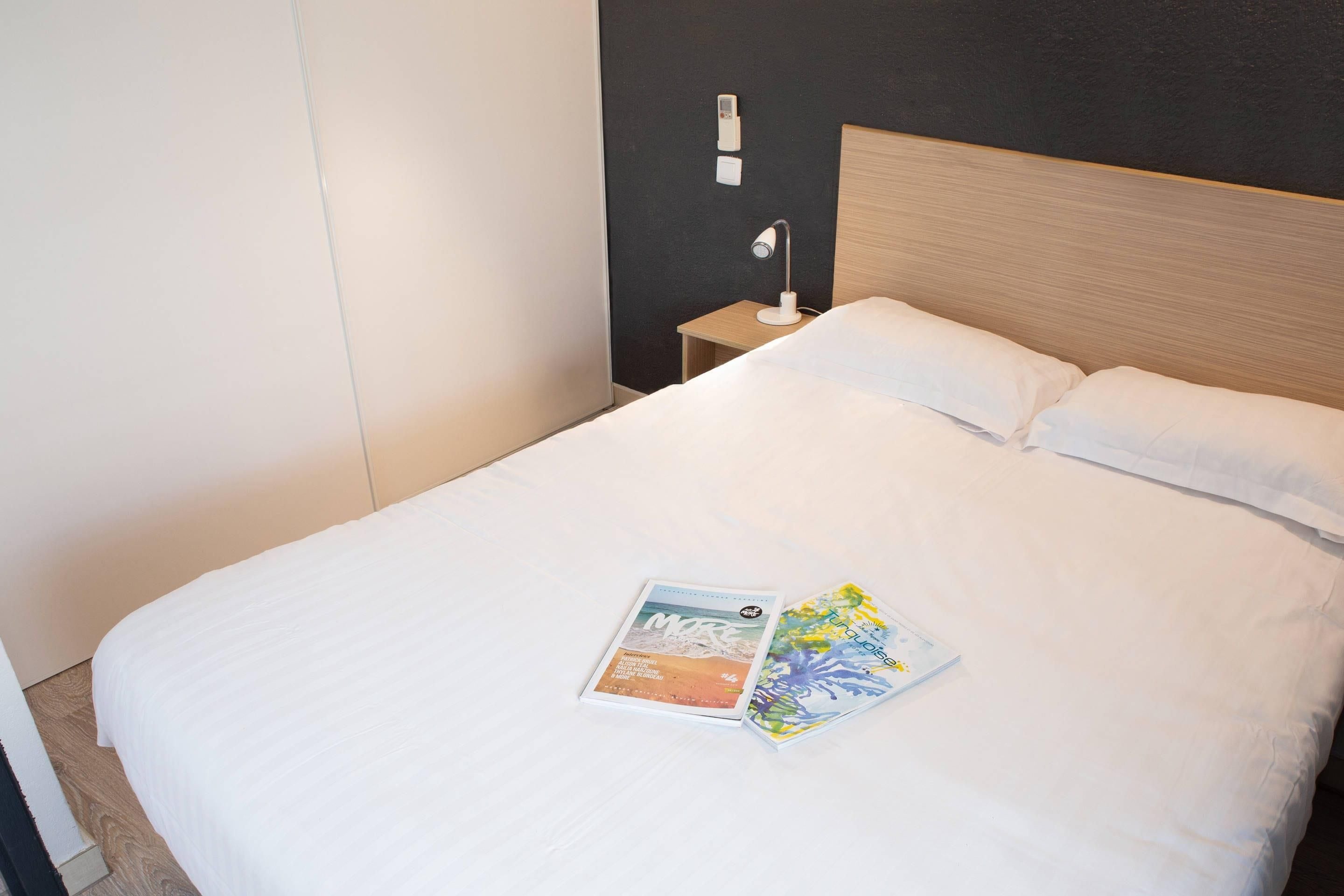 A neatly made bed with white linens, a bedside lamp, and two magazines on the bed.