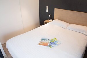 Una cama bien tendida con sábanas blancas, una lámpara de noche y dos revistas sobre la cama.