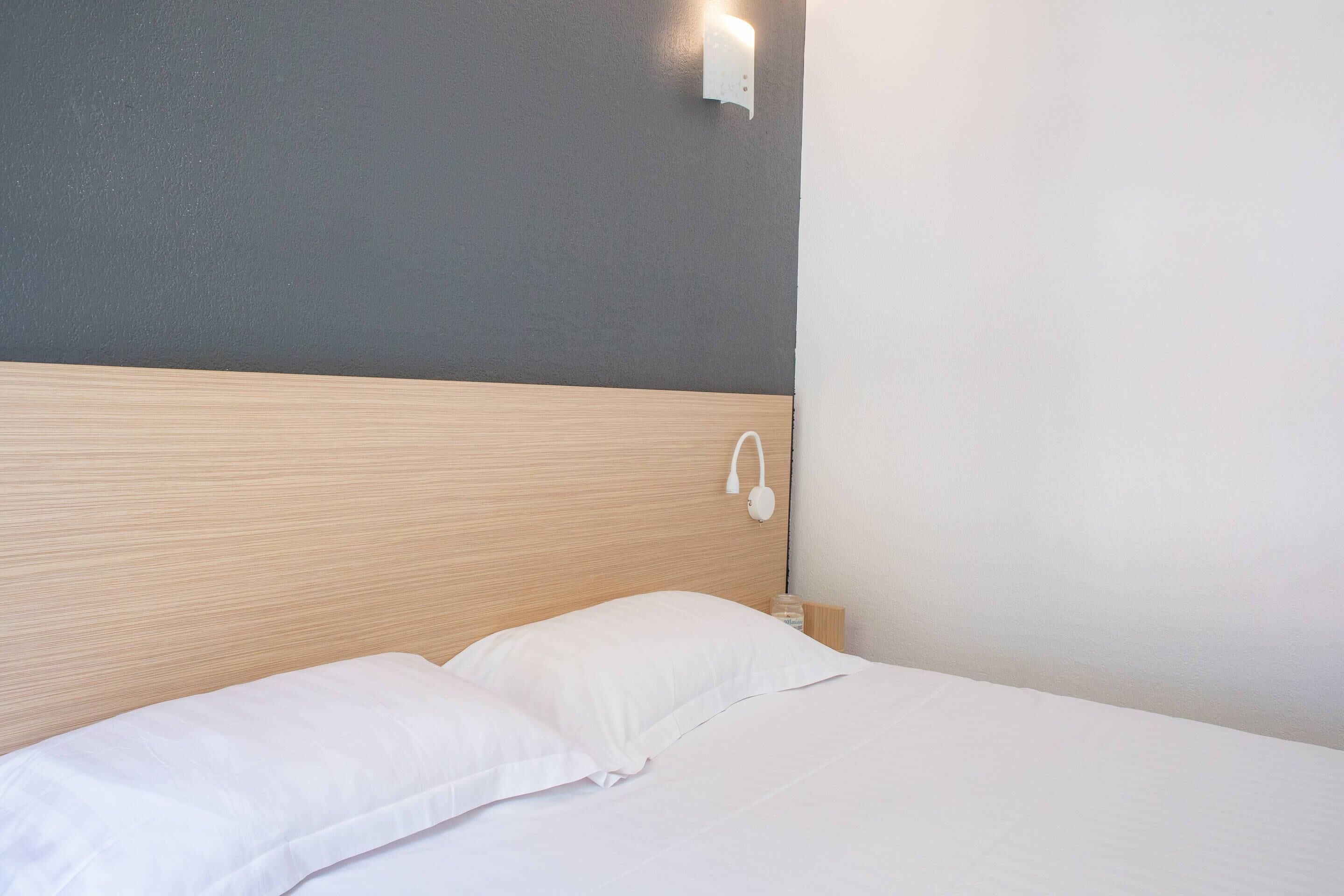 Ein ordentlich bezogenes Bett mit weißen Bettwäsche, einem Holzkopfteil und einer Wandleuchte.