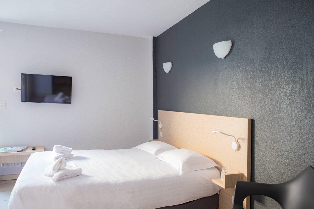 A hotel room with a bed, a TV, a desk with a book, and a chair.
