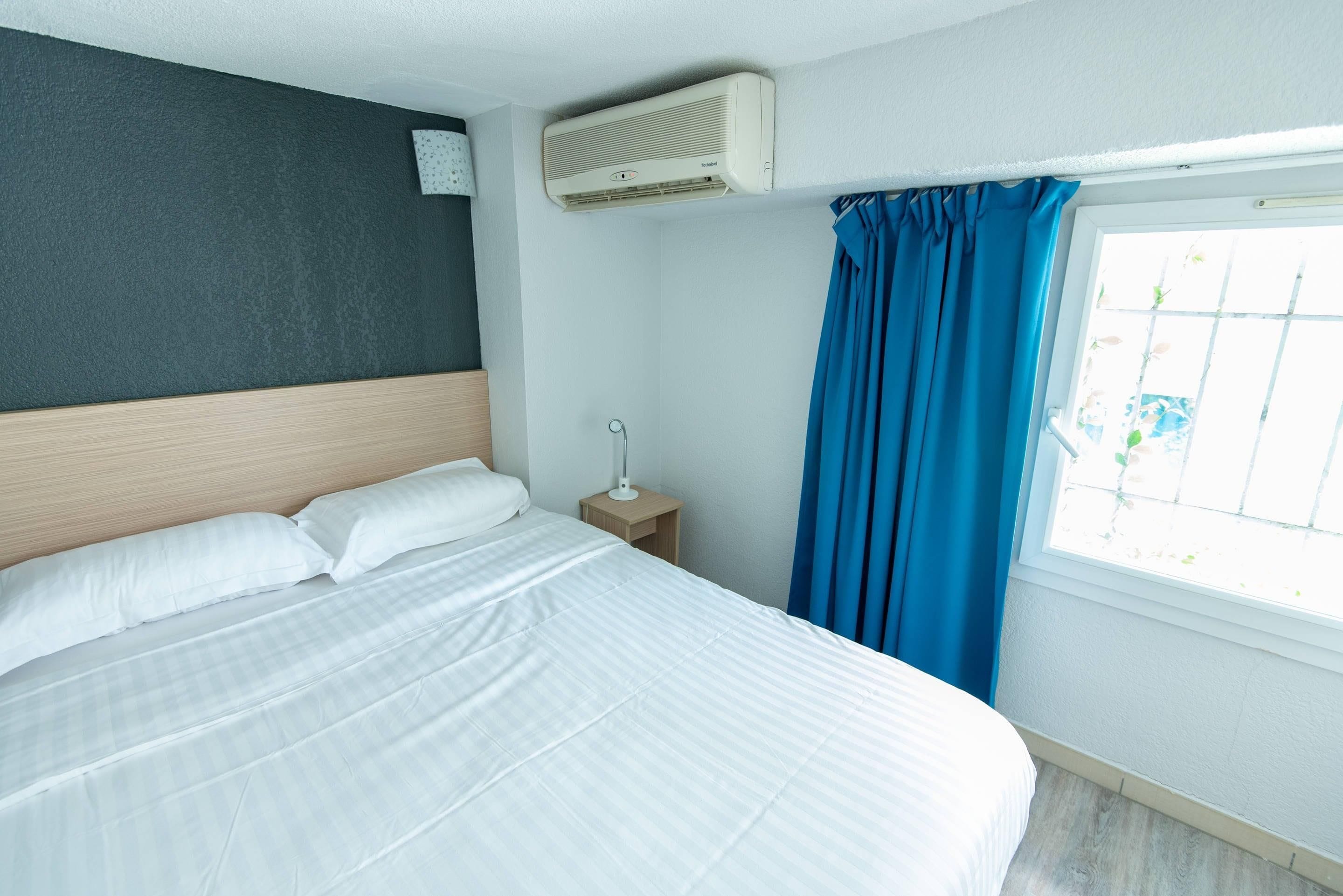 Quarto de hotel com uma cama, uma mesinha de cabeceira, uma janela com cortinas e um aparelho de ar condicionado.