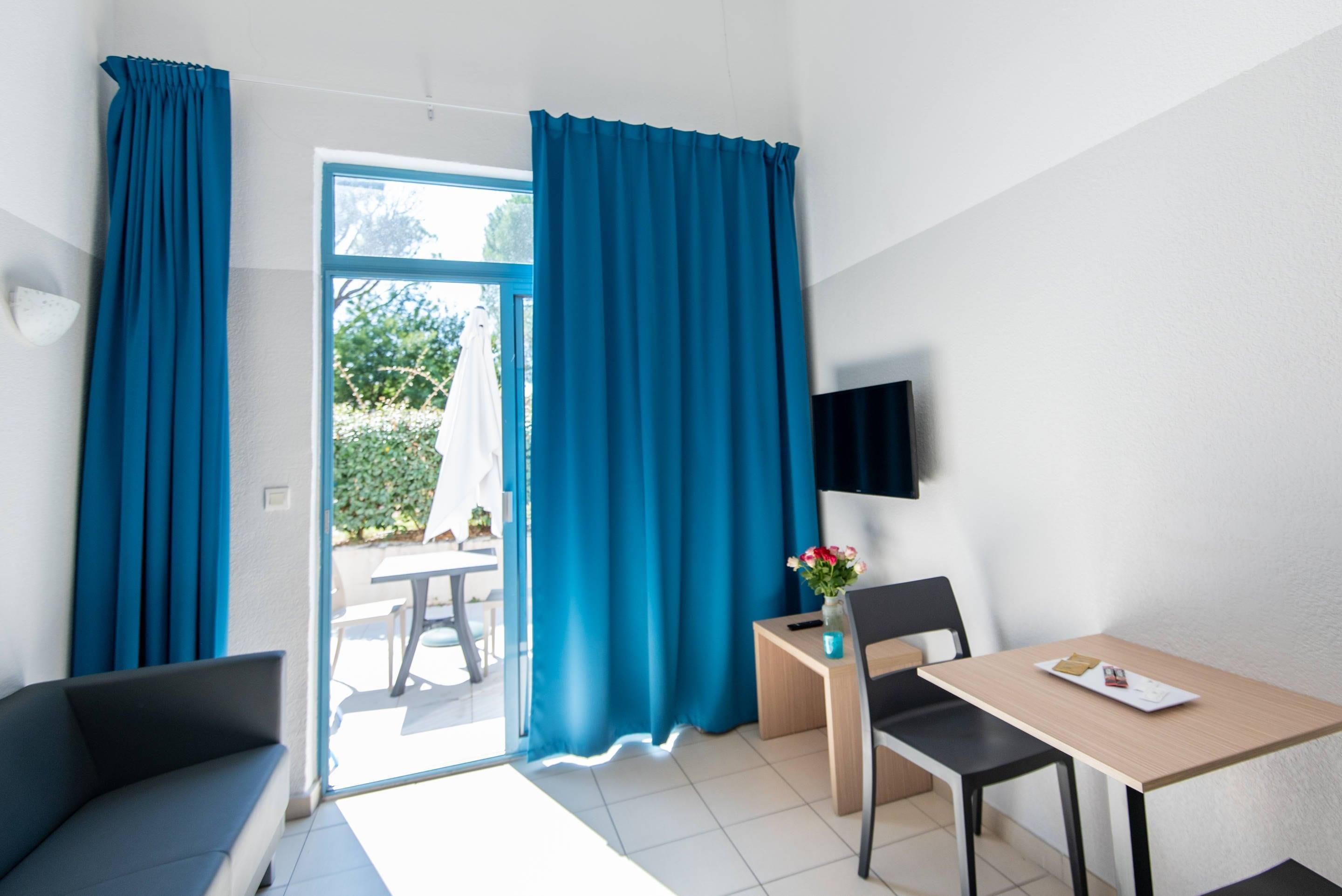 Habitació amb sofà, taula i una cadira, televisió penjada a la paret i finestra amb cortines.