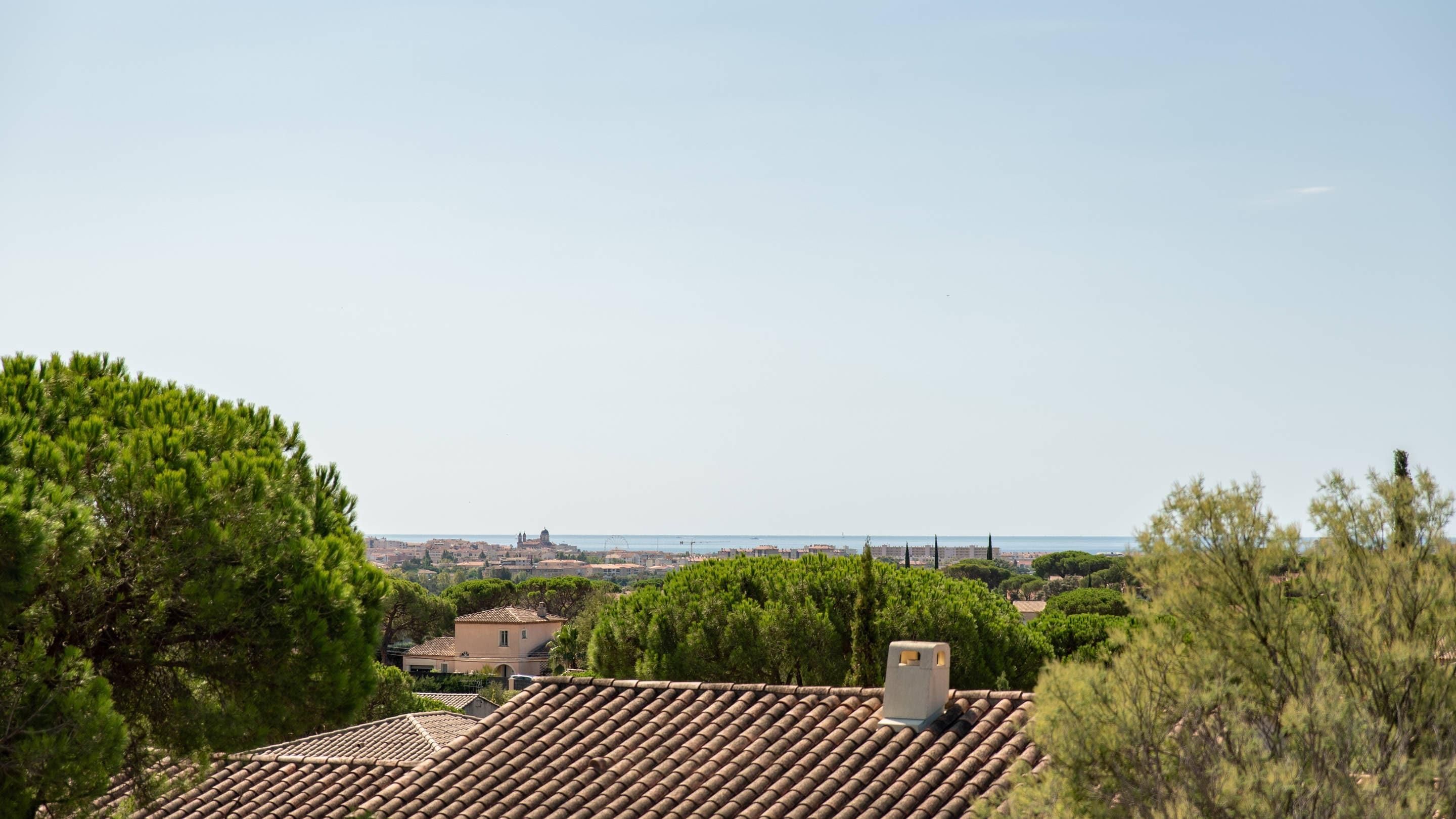 Zona residencial con tejados de tejas rojas, abundante vegetación y una vista lejana de un paisaje urbano costero.