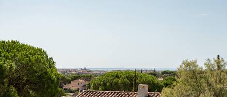 A residential area with terracotta-tiled roofs, dense greenery, and a distant view of a coastal cityscape.