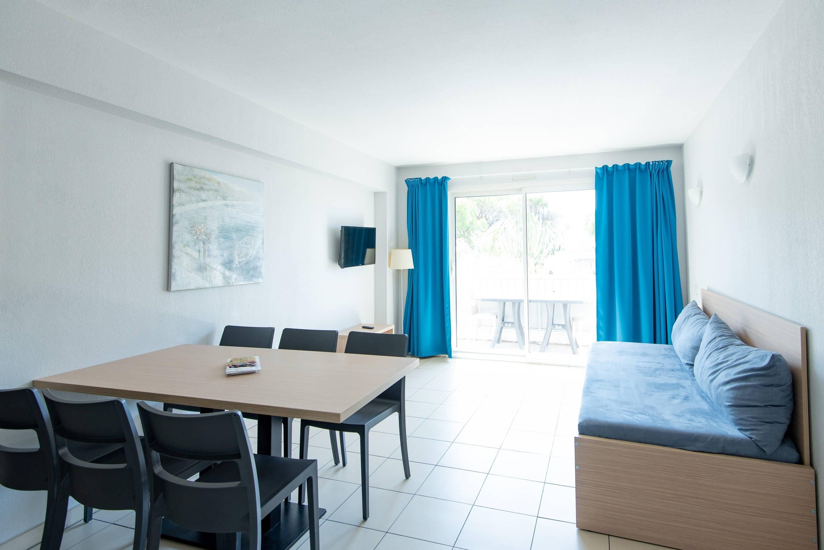 Une pièce comprenant un lit, une table à manger avec des chaises, un téléviseur et une fenêtre avec des rideaux.