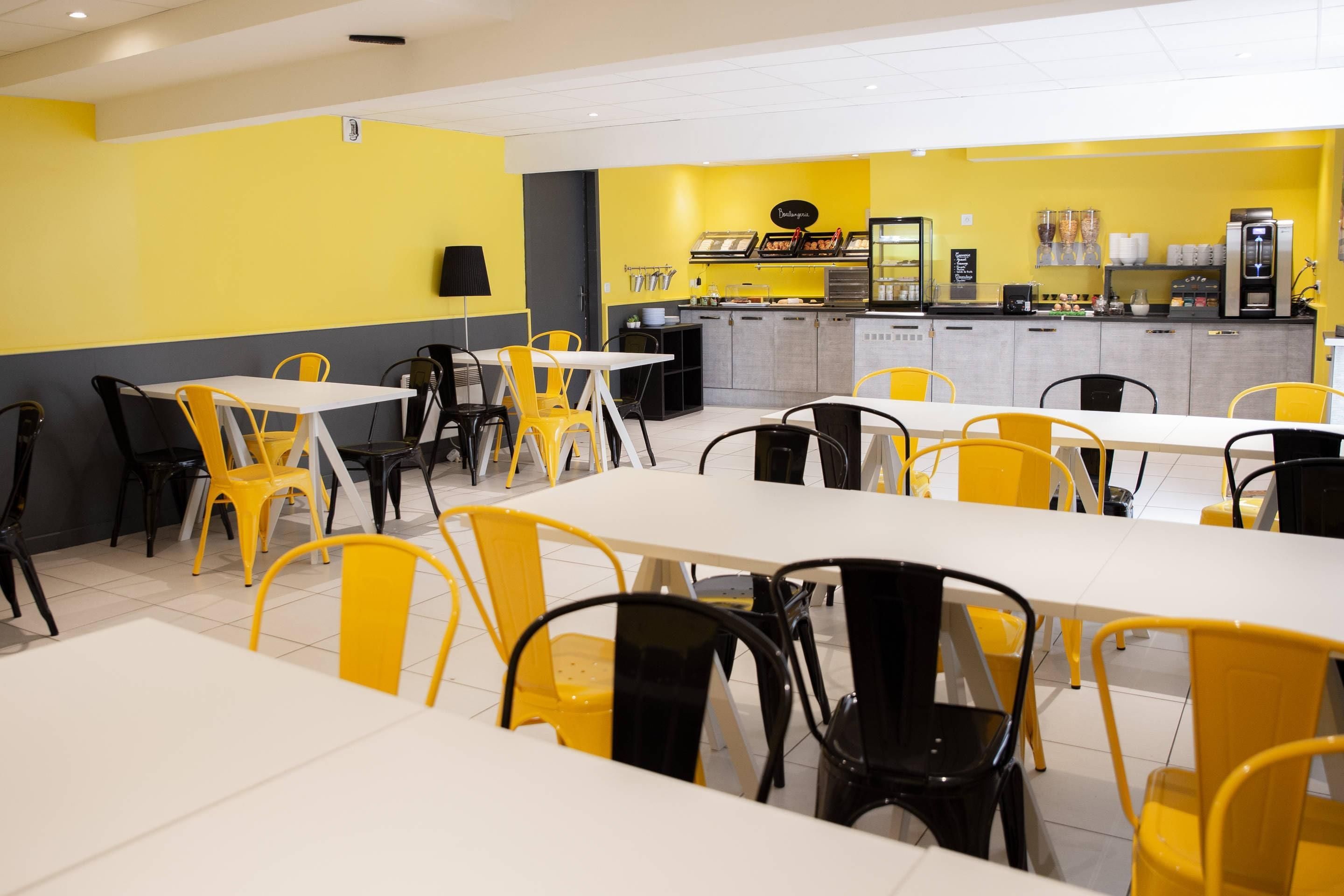 Un restaurante con sillas amarillas y negras, mesas blancas y una barra con exhibición de alimentos.