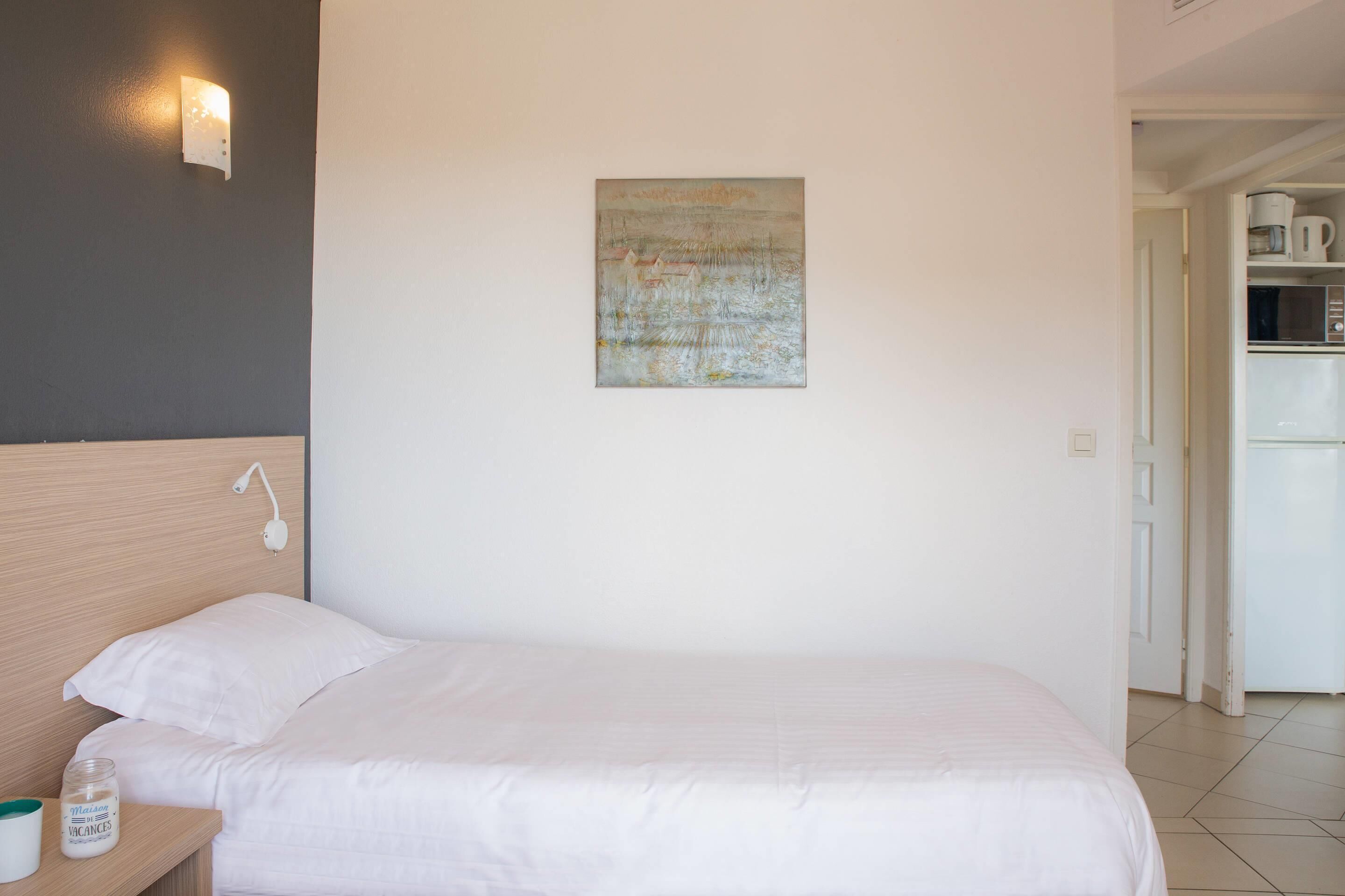 A hotel room with a bed, a nightstand, a wall-mounted lamp, a painting, and a kitchenette in the background.