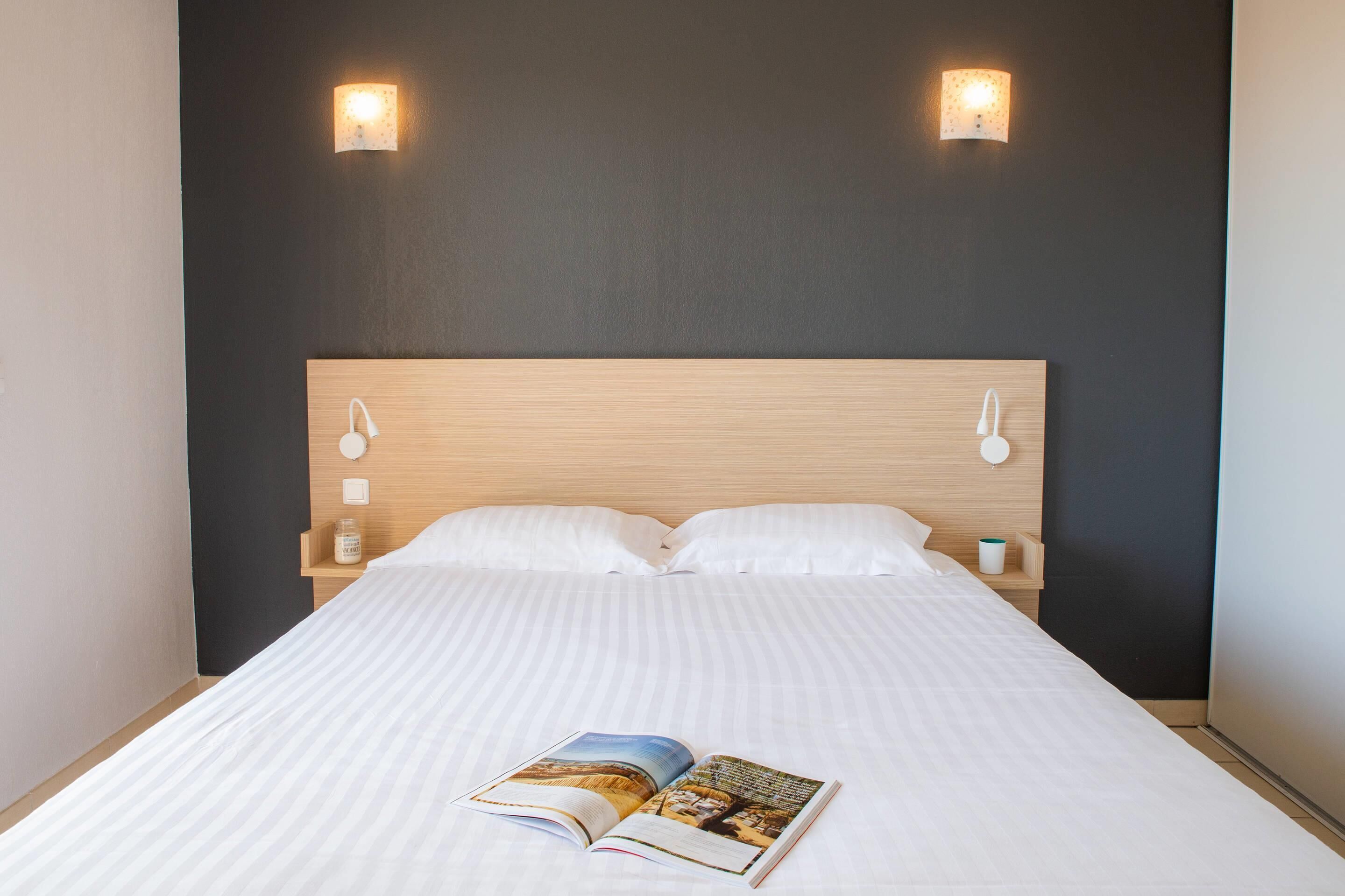 Ein ordentlich bezogenes Bett mit einem Buch darauf, zwei Wandleuchten und einem hölzernen Kopfteil.
