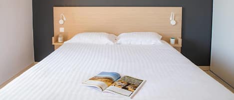 Cama bem arrumada com um livro, duas luzes na parede e cabeceira de madeira.