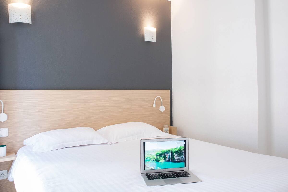 A hotel room with a bed, a laptop on the bed, a nightstand with a lamp, and a wall-mounted light fixture.