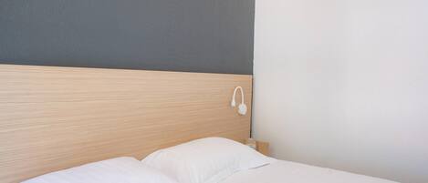Una cama bien tendida con sábanas blancas, un cabecero de madera y una lámpara fijada a la pared.