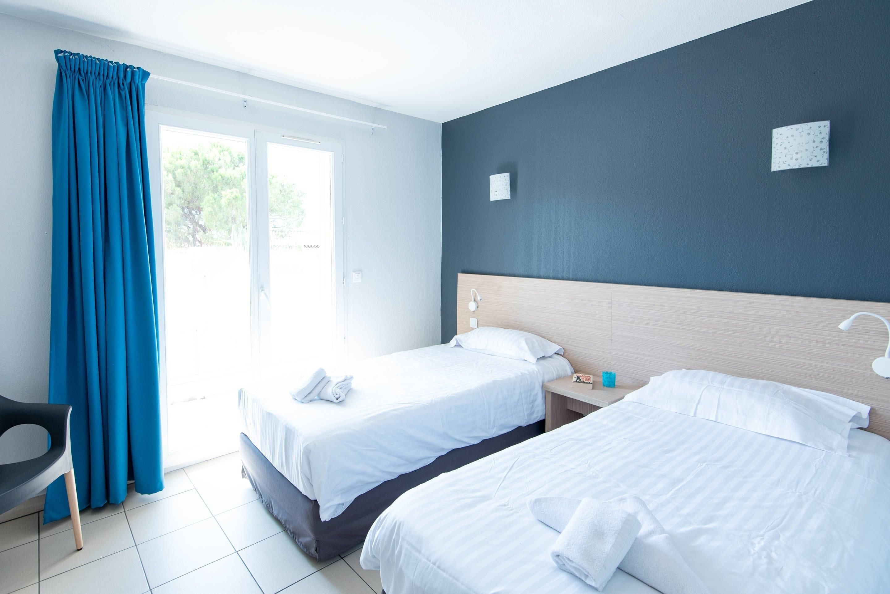 Hotellihuone, jossa on kaksi sänkyä, työpöytä, tuoli ja suuri ikkuna, jossa on verhot.