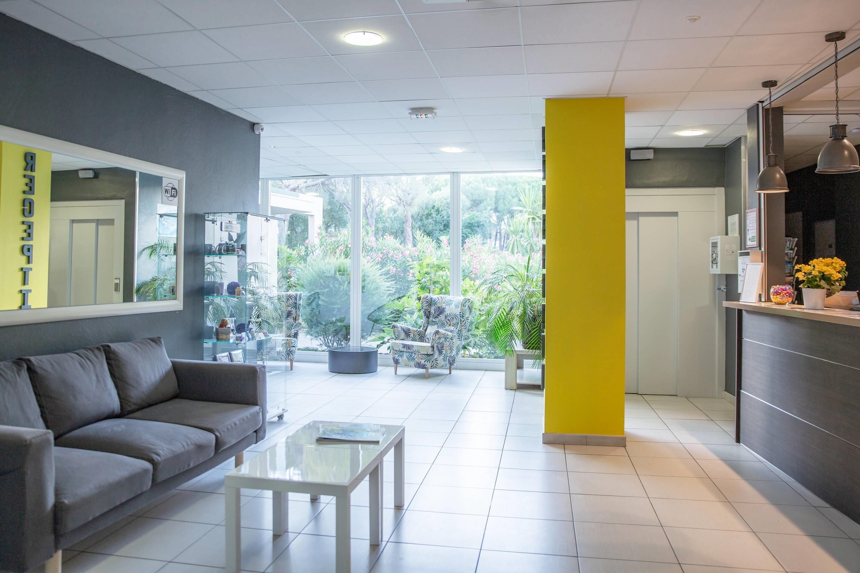 Un hall d’hôtel moderne avec un canapé gris, une table basse en verre et un pilier jaune.