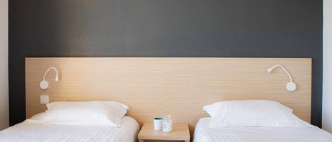 Habitación de hotel con dos camas, cabecera de madera, lámparas de noche y mesita de noche con una botella y un vaso.