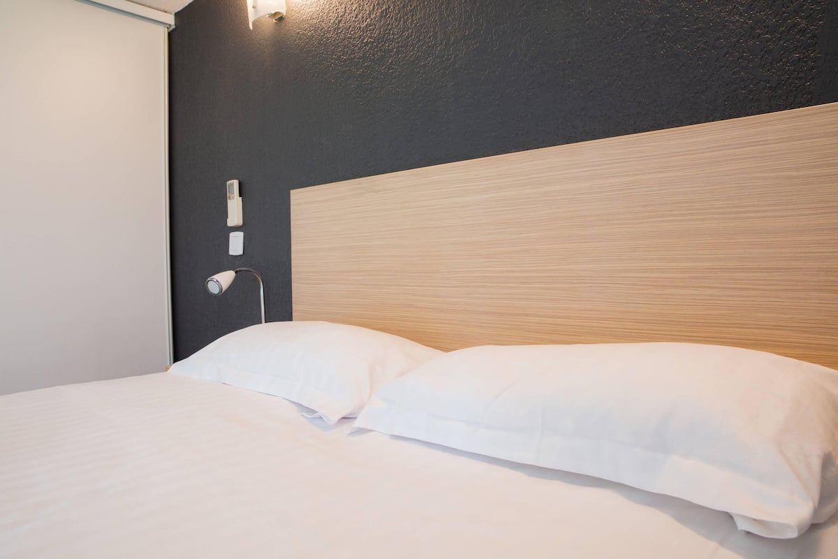 A hotel room with a wooden headboard, white bedding, and a wall-mounted light fixture.