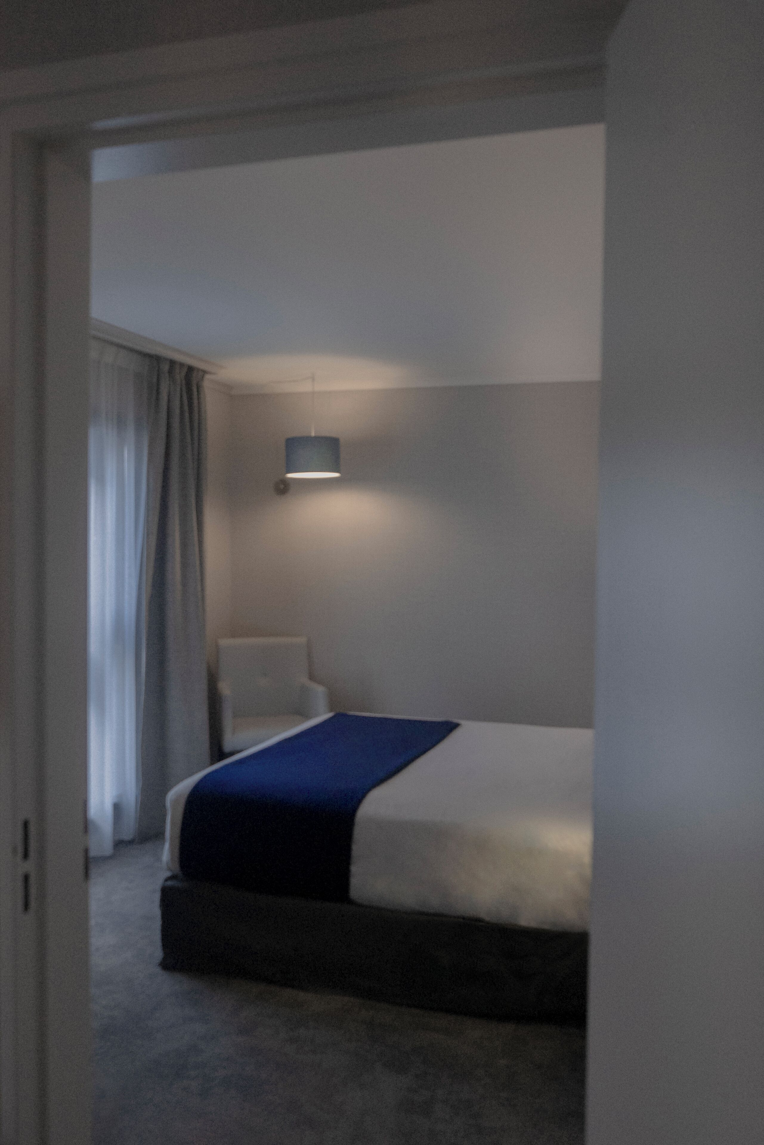 Una camera d'albergo con un letto, una sedia, una lampada e una finestra con tende.