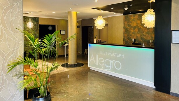 A hotel reception area with a modern design, featuring a reception desk labeled "Best Western Allegro," ambient lighting, and potted plants.