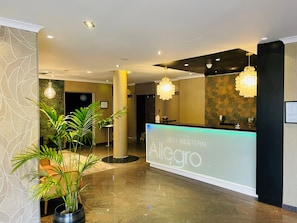 Zona de recepción de hotel con un diseño moderno, que incluye un mostrador de recepción con la etiqueta "Best Western Allegro", iluminación ambiental y plantas en macetas.